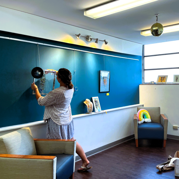 A white woman with her back turned to camera, facing a green wall where she is hanging a piece of art.