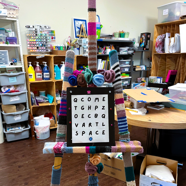 an easel with a black and white sign on it in the foreground, and a filled art supply room in the background.